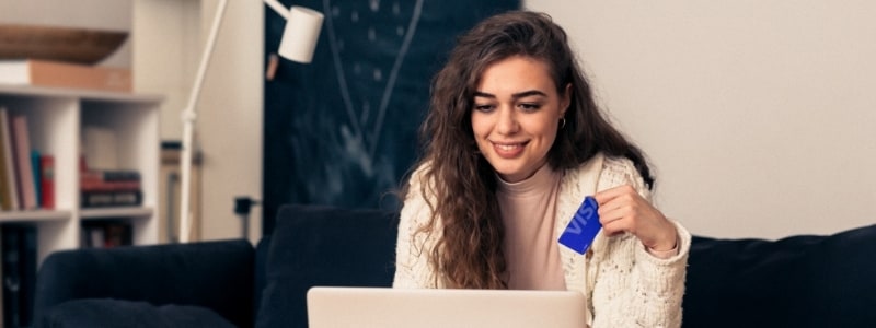 Young woman at home, smiling while using a laptop and holding a blue Visa card
