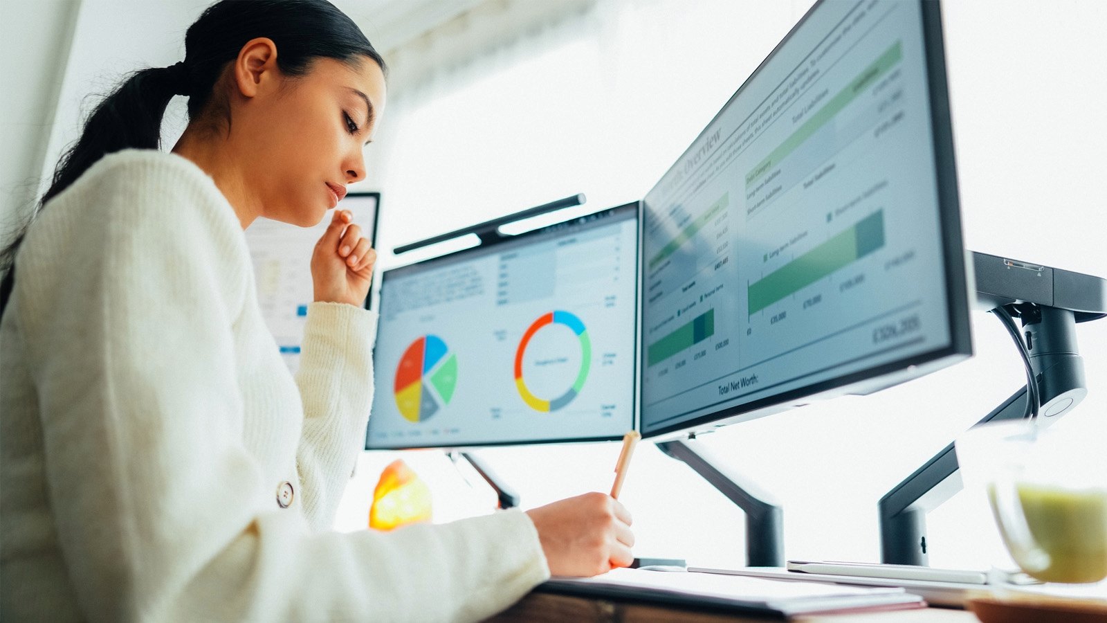 Woman sitting at desk working with multiple monitors in front of her showing pie charts and other data visualizations