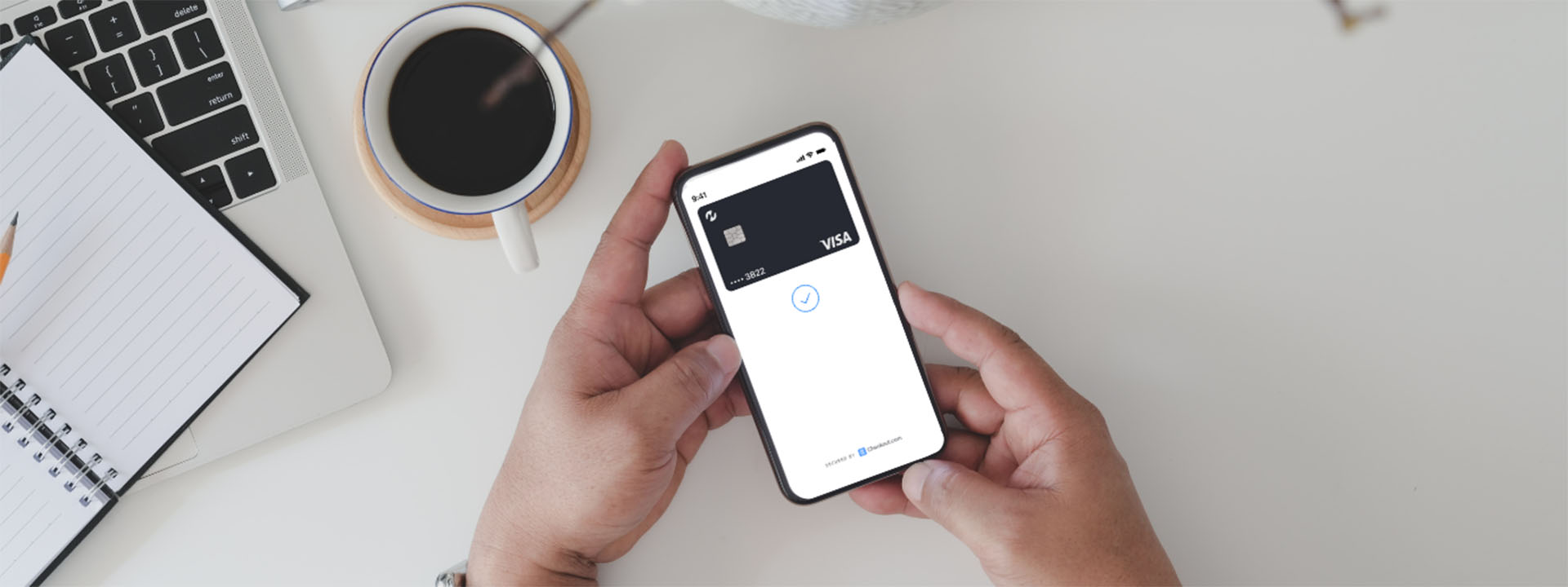 Person holding a smartphone displaying a Visa card that is powered by checkout.com in a digital wallet app, with a laptop, notebook, coffee cup, and camera on a white desk in the background.