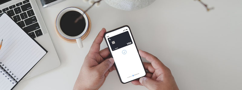 Person holding a smartphone displaying a Visa card that is powered by checkout.com in a digital wallet app, with a laptop, notebook, coffee cup, and camera on a white desk in the background.