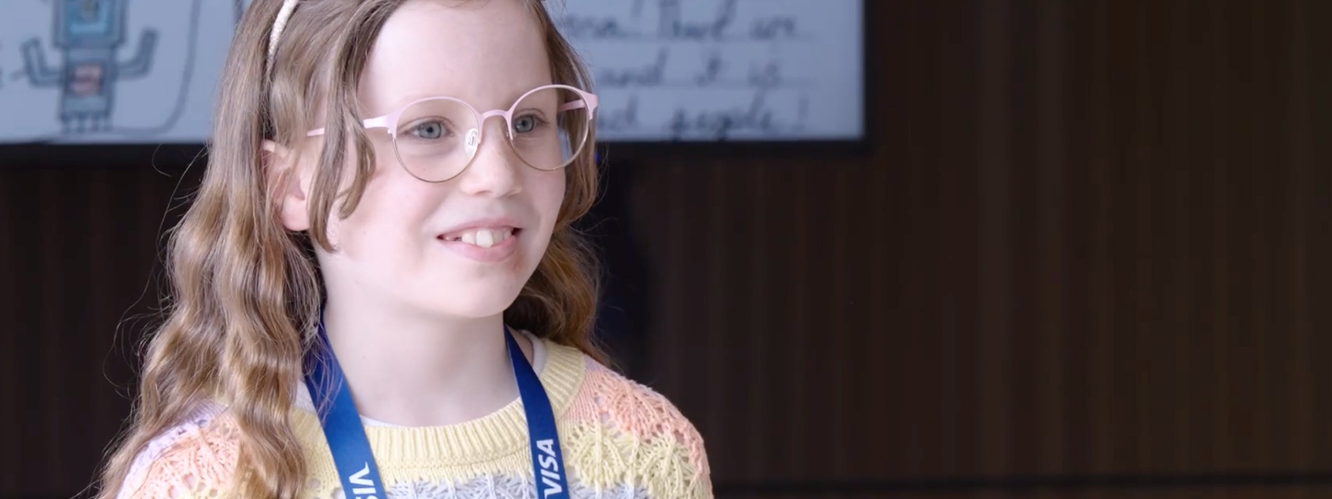 A young girl smiling wearing pink glasses and a Visa lanyard around her neck