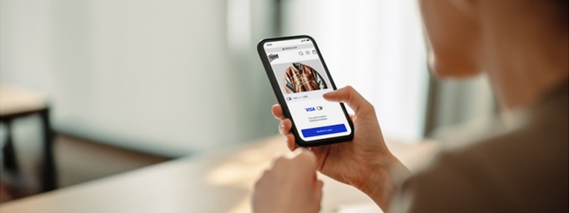 Hand holding phone that on screen shows an in progress online clothing purchase using Visa’s Click to Pay payment option.