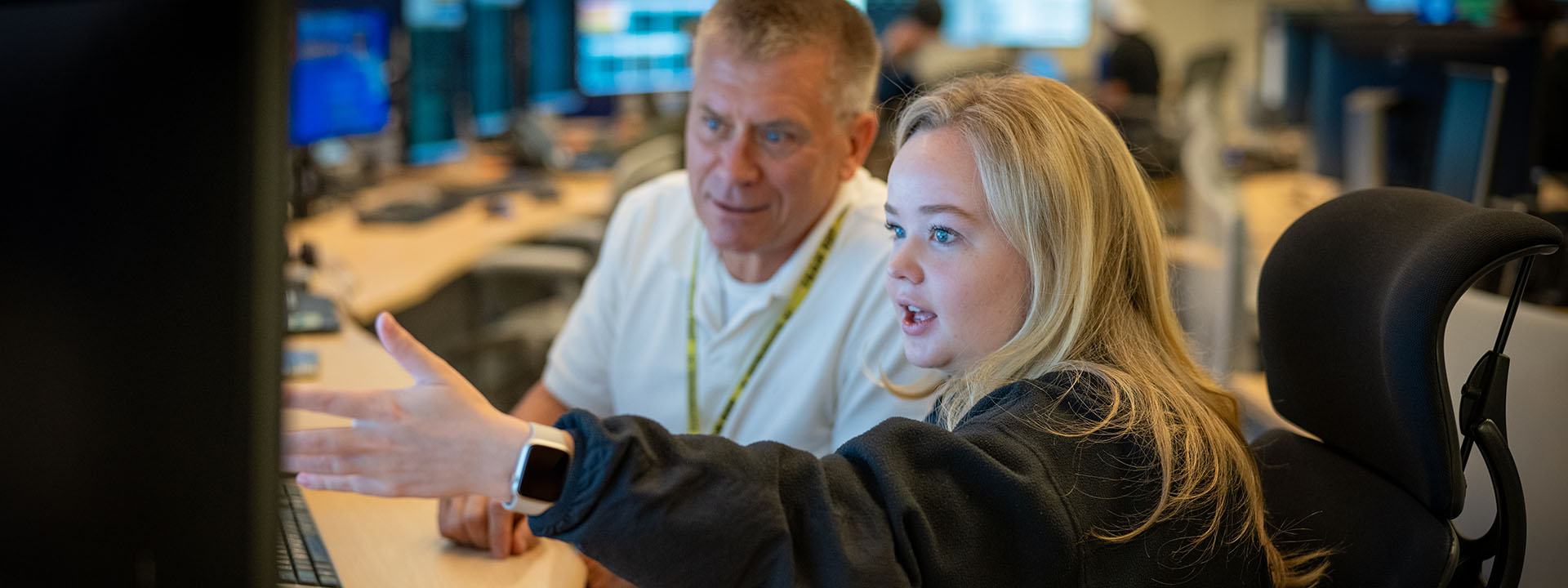 In a control room, a man in a gray/beige shirt and a blonde woman in a black sweatshirt focus on computer screens, surrounded by technical equipment.