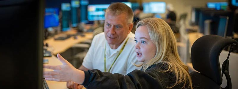 In a control room, a man in a gray/beige shirt and a blonde woman in a black sweatshirt focus on computer screens, surrounded by technical equipment.