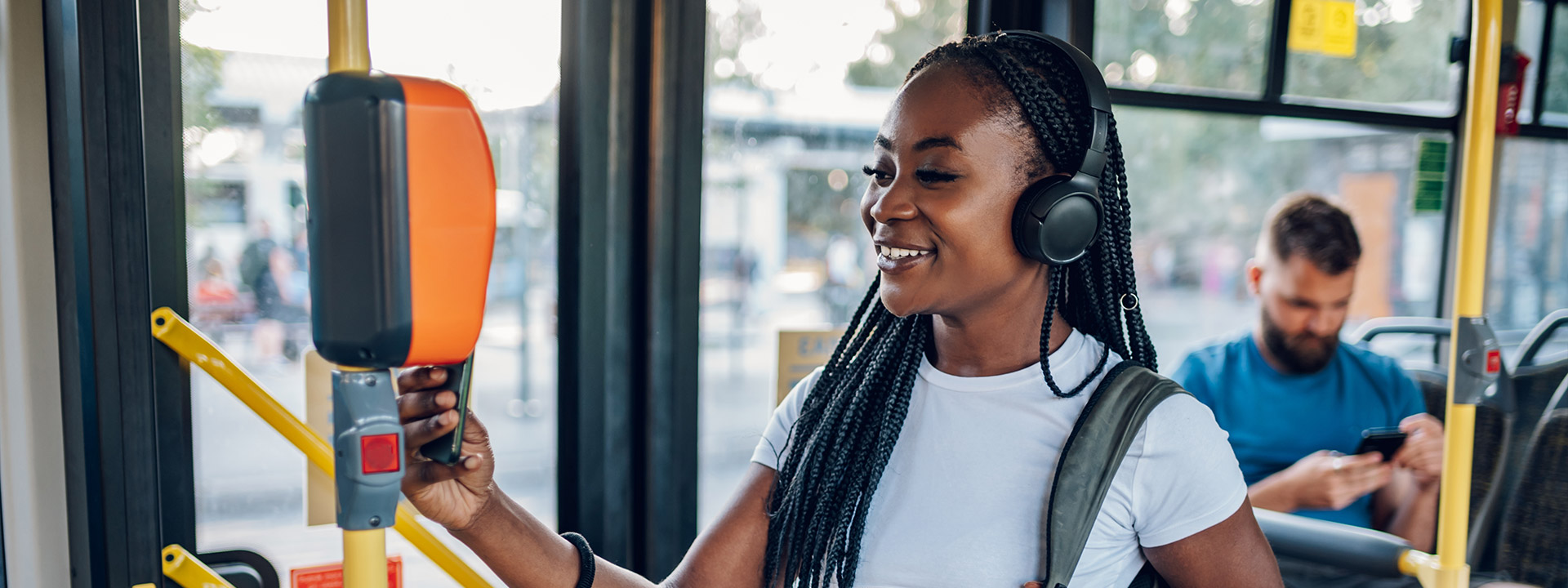 A woman, smiling and wearing headphones, taps her card to pay her fair on a city bus