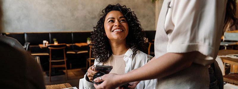Woman using smartphone while paying restaurant bill with contactless payment during dinner party with friends