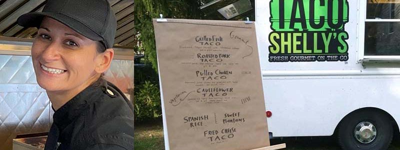 A smiling woman in a black chef's coat stands by "TACO SHELLY'S" food truck, showcasing a menu of delicious taco options.