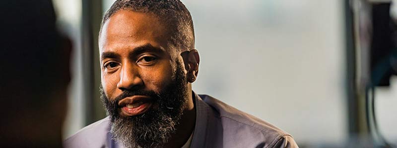 A dark-skinned man with a beard and dreadlocks in a bun, wearing a gray collared shirt, engaged in conversation. A blurred background suggests an interview setting.