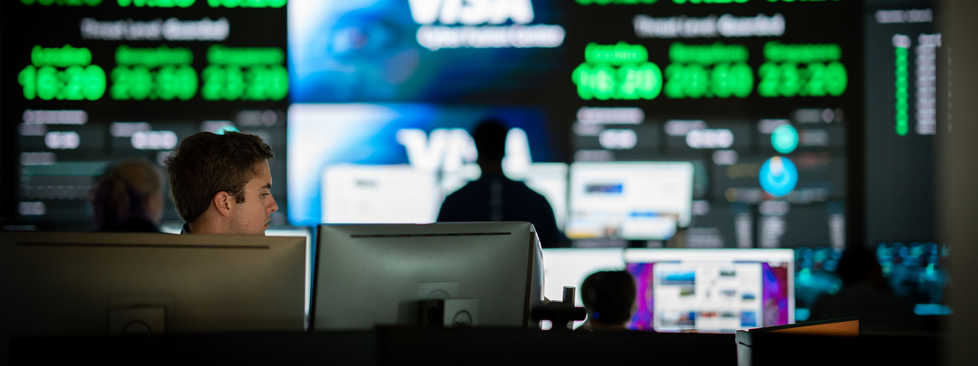 Visa employee working behind two computers in front of a wall of screens.
