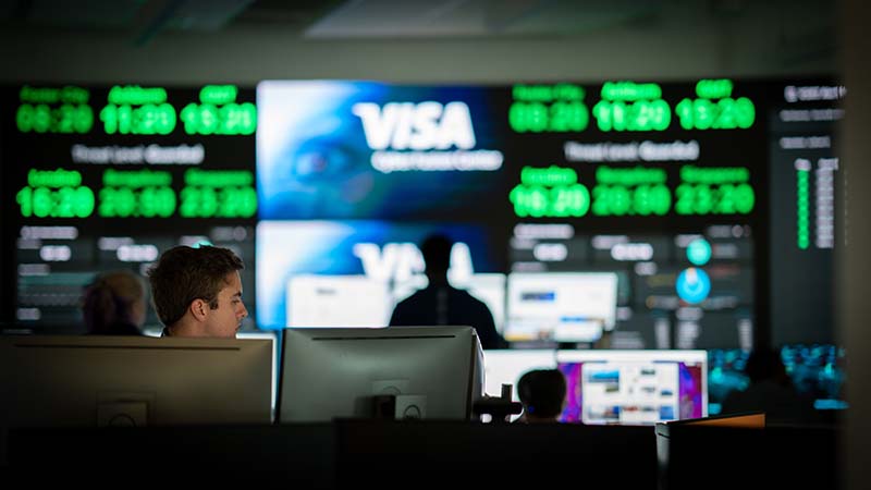 Visa employee working behind two computers in front of a wall of screens.