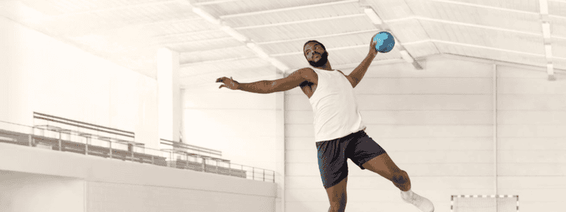 Handballer Dika Mem holding a handball mid-air while jumping in a white gymnasium.