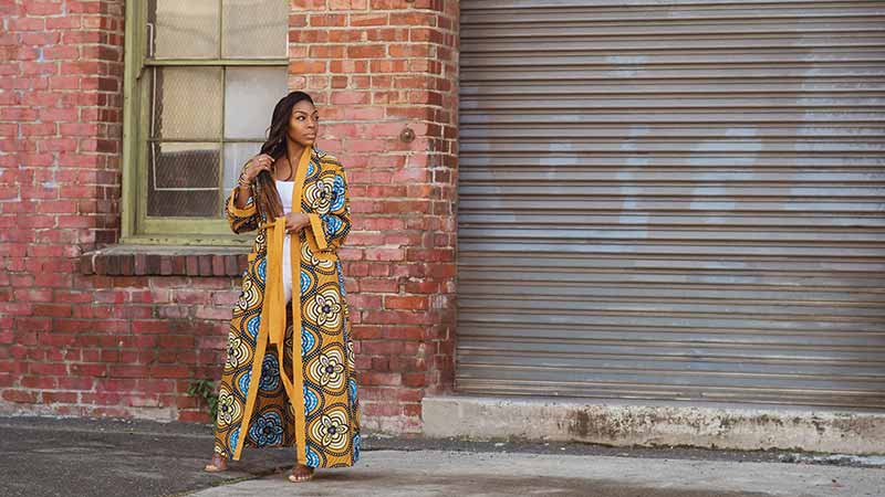 Love Iguehi founder Iguehi James wears her ankle-length Fula Duster in front of a closed corrugated steel garage door.