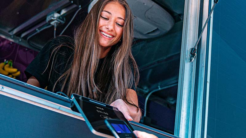 A woman pops out of a food truck window holding out her tablet to receive payment from someone with a mobile phone.