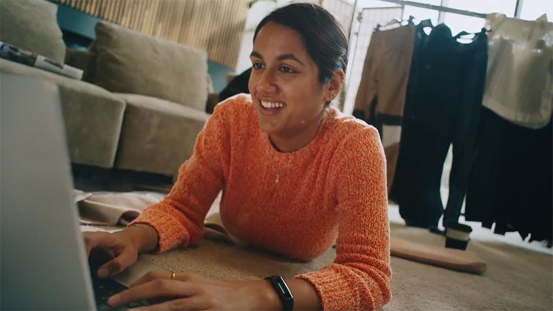 A woman in an orange sweater lies on the floor typing on a laptop while smiling