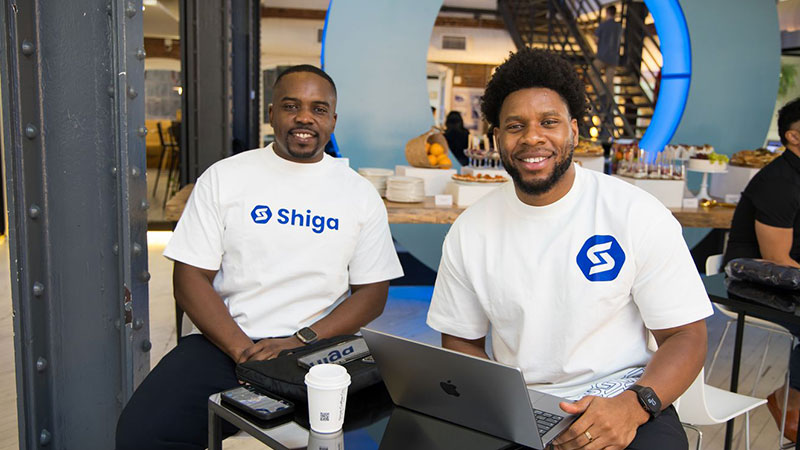 Two men smiling at a table with laptops and coffee, wearing white Shiga t-shirts. Background features a modern café with lit blue accents.