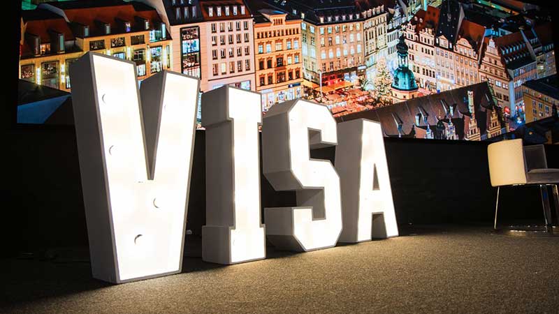 Lit letters spelling "VISA" stand on a dark stage with a colorful cityscape backdrop. The scene conveys a modern, vibrant, and dynamic tone.