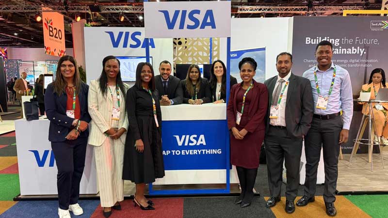 A diverse group of nine professionally-dressed people smiles in front of a Visa booth at a vibrant trade show, creating an optimistic and energetic atmosphere.