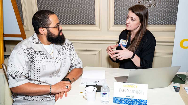 A man and woman engage in conversation at a table with a laptop and a "CONNECT BRANDING" sign during a business event.