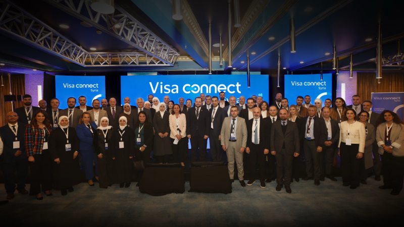 Group photo of diverse adults in business attire at a conference, standing in front of a blue "Visa Connect" backdrop. The atmosphere is professional and collaborative.
