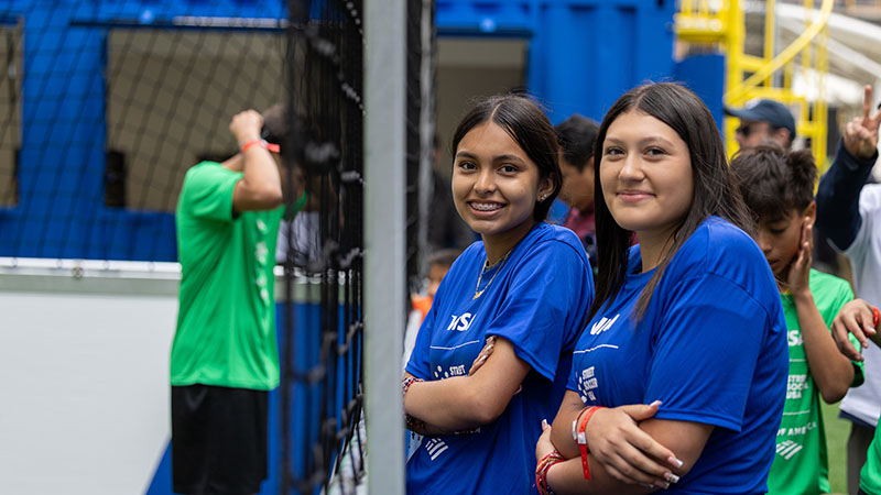 Two young soccer players wearing blue shirts stand next to the street soccer field, looking at the camera.